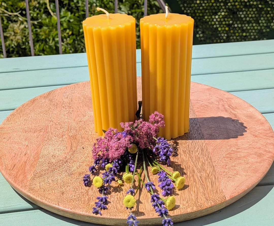 Zwei große Bienenwachsstumpen mit feinen Längsrillen auf Holzscheibe mit Trockenblumen
